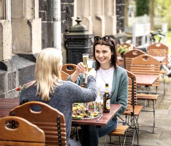 Zwei Frauen sitzen an einem Holztisch im Freien und stoßen mit Getränken an. Im Hintergrund sind Fahrräder zu sehen., © Eifel Tourismus GmbH, Dominik Ketz Zwei Frauen sitzen an einem Holztisch im Freien und stoßen mit Getränken an. Im Hintergrund sind Fahrräder zu sehen., © Eifel Tourismus GmbH, Dominik Ketz