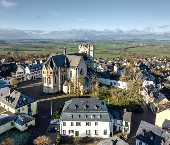 Luftaufnahme von Münstermaifeld mit der Stiftskirche, umgeben von Häusern. Im Hintergrund weite Felder und Windräder unter blauem Himmel., © Eifel Tourismus GmbH, D. Ketz Luftaufnahme von Münstermaifeld mit der Stiftskirche, umgeben von Häusern. Im Hintergrund weite Felder und Windräder unter blauem Himmel., © Eifel Tourismus GmbH, D. Ketz