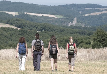 Vier Wanderer mit Rucksäcken gehen über ein Feld. Im Hintergrund ist die Burgruine Monreal zu sehen, umgeben von bewaldeten Hügeln., © Traumpfade/Kappest Vier Wanderer mit Rucksäcken gehen über ein Feld. Im Hintergrund ist die Burgruine Monreal zu sehen, umgeben von bewaldeten Hügeln., © Traumpfade/Kappest