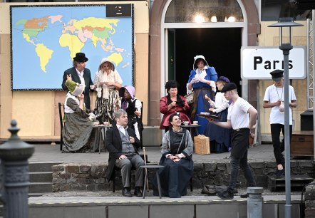 Der Theaterverein TheaterKultur Kaisersesch bei der Aufführung von 'In 80 Tagen um die Welt', © TheaterKultur Kaisersesch e. V., Christoph Gerhartz Mitglieder des Theaterverein Kaisersesch spielen auf einer Freilichtbühne eine Szene aus „In 80 Tagen um die Welt“: In historischen Kostümen sitzen und stehen mehrere Darsteller vor einer Weltkarte und einem „Paris“-Schild, während ein Kellner Getränke serviert und weitere Figuren im Hintergrund Tee trinken.