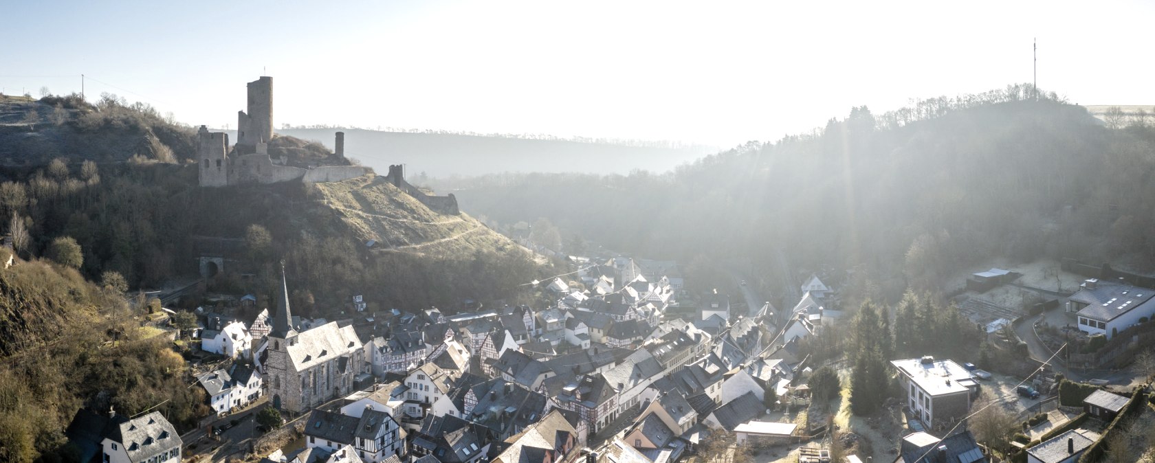 Luftaufnahme von Monreal, einem malerischen Dorf mit Fachwerkhäusern und einer Burgruine auf einem Hügel im Hintergrund, umgeben von bewaldeten Hügeln., © Eifel Tourismus GmbH, D. Ketz