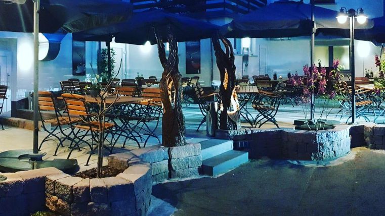 Ein gemütlicher Außenbereich eines Restaurants mit mehreren Tischen und Stühlen. Über den Tischen sind Sonnenschirme und schöne Pflanzen sind dekorativ platziert.