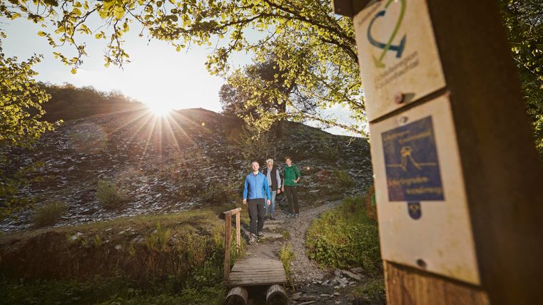 Twee personen wandelen op een smal pad over een kleine brug. Op de achtergrond schijnt de zon door de bomen.
