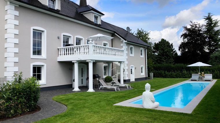 Ein elegantes Haus mit einem gepflegten Garten und einem Pool. Im Hintergrund strahlt der Himmel mit einigen Wolken.