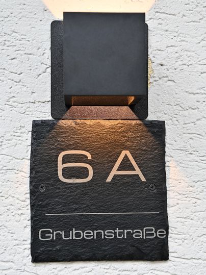 Eine moderne Hausnummer 6A aus schwarzem Stein mit dem Schriftzug "Grubenstraße". Sie ist an einer weißen Wand montiert und wird von einer Deckenleuchte beleuchtet.