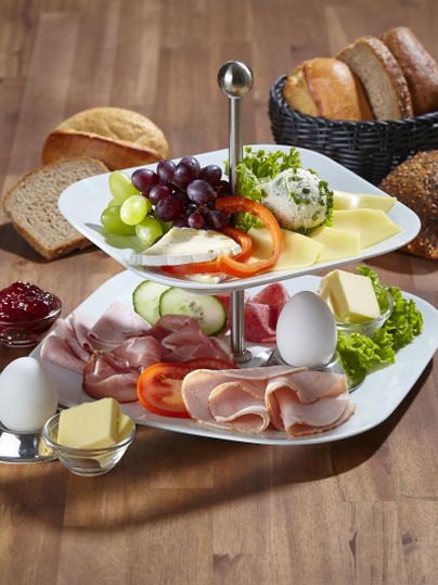 Ein attraktives zweistöckiges Frühstücksbrett mit Käse, Wurst, frischem Obst und Gemüse. Im Hintergrund befinden sich verschiedene Brotsorten und Marmelade.