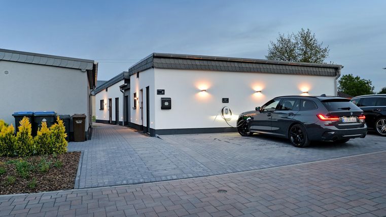 Ein modernes Haus mit hellem Außenanstrich und einer Auffahrt. Ein schwarzes Auto steht geparkt, umgeben von grünen Pflanzen.