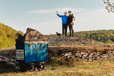 Zwei Wanderer mit Hund machen ein Selfie auf einem Schieferfeld in der Eifel. Links im Vordergrund steht ein blauer Förderwagen mit Schiefergestein, dahinter Wälder und grünes Hügelland unter blauem Himmel.