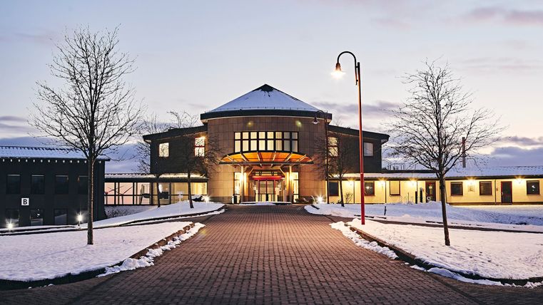 Ein modernes Gebäude mit einer einladenden Eingangstür und großen Fenstern. Der Weg ist von Schnee und Bäumen gesäumt, und die Dämmerung verleiht der Szene eine besondere Stimmung.