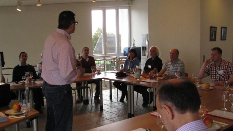 Een man staat voor in een conferentiezaal en spreekt tot een groep luisteraars. De aanwezigen zitten aan een lange tafel met drankjes en noteren mogelijk belangrijke informatie.