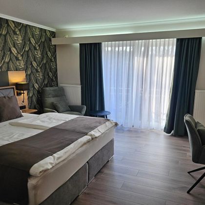 Ein modernes Hotelzimmer mit einem großen Bett und stilvoller Einrichtung. Viel Tageslicht strömt durch die Fenster, und ein bequemer Sessel steht zur Verfügung.