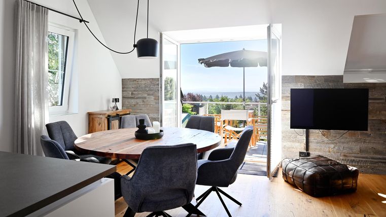 Ein modernes Esszimmer mit einem runden Holztisch und bequemen Stühlen. Große Fenster bieten einen Blick auf die Terrasse und die Landschaft.