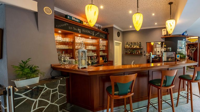 Eine gemütliche Bar mit einem langen Holztheken, modernen Stühlen und warmem Licht. Im Hintergrund sind Gläser und eine gut bestückte Bar zu sehen.