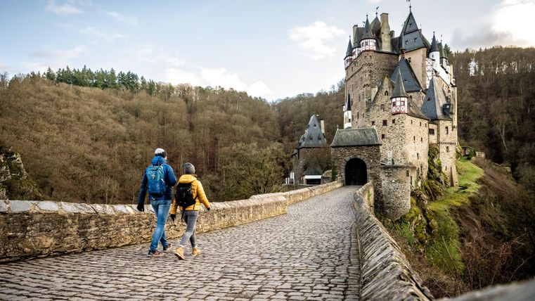 Ein Paar geht über eine steinerne Brücke zu einer beeindruckenden Burg. Die Umgebung ist grün und bewaldet, und der Himmel hat eine leichte Bewölkung.