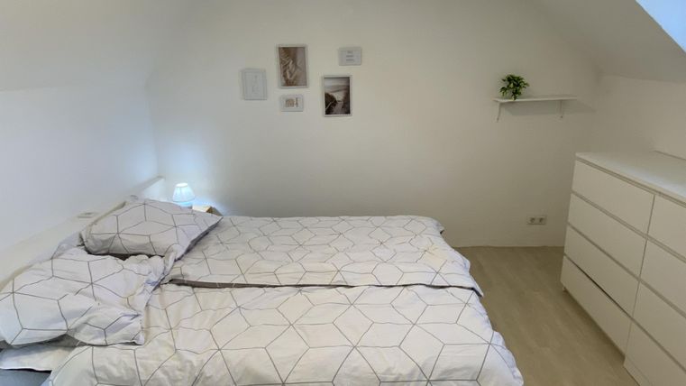 Ein gemütliches Schlafzimmer mit einem Bett und weißer Bettwäsche. An der Wand hängen einige Bilder, und es gibt eine kleine Regale mit einer Pflanze.