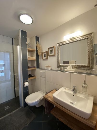 Ein modernes Badezimmer mit einer Dusche, einem Waschbecken und eleganten Dekorationselementen. Die Wände sind hell gefliest, und ein großer Spiegel fügt einen stilvollen Akzent hinzu.