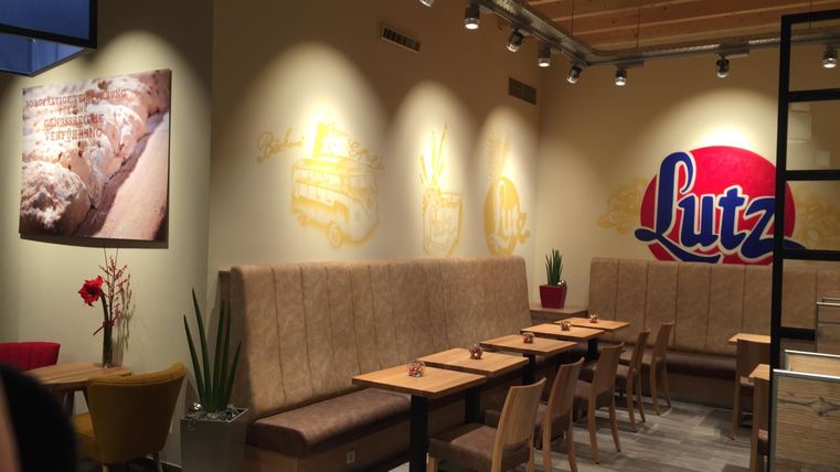 Ein modernes Café mit holzverkleideten Wänden und gemütlichen Sitzplätzen. An der Wand ist das Logo "Lutz" sowie ein rotes Bild zu sehen.