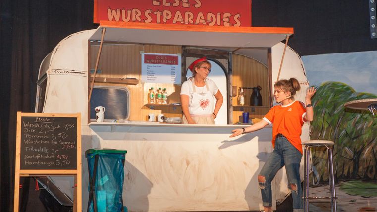 Zwei Schauspieler des Theatervereins Düngenheim stehen an einem Essensstand und unterhalten sich.