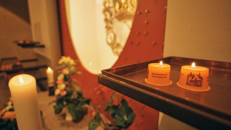 Zwei brennende Kerzen auf einem Tisch in einer Innenansicht. Im Hintergrund sind Blumen und eine religiöse Wandfläche sichtbar.