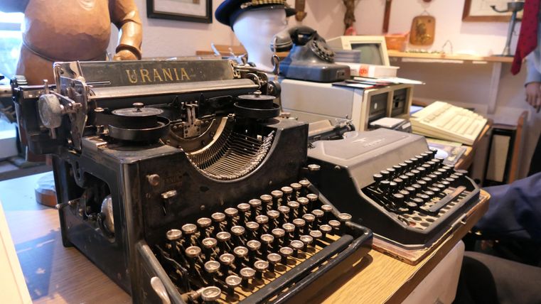 Eine alte Schreibmaschine namens Urania steht auf einem Tisch. Daneben befindet sich eine weitere Schreibmaschine in dunklem Design.