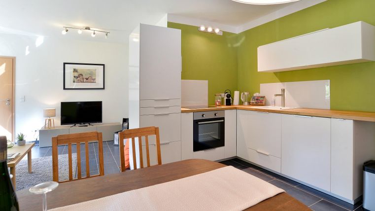 Eine moderne Küche mit grünen Wänden und einer hellen, minimalistischen Einrichtung. Der Essbereich hat einen Holztisch mit Stühlen und einem Blick auf einen Fernseher.