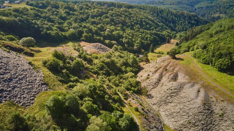 Eine grüne Landschaft mit Hügeln und bewaldeten Flächen. Im Vordergrund sind Steinhaufen und unebenes Gelände sichtbar.