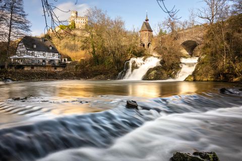 Ein ruhiger Fluss mit sanften Wasserfällen fließt vor einer alten Burg. Umgeben von Bäumen und einem historischen Gebäude ist die Szene idyllisch und malerisch.