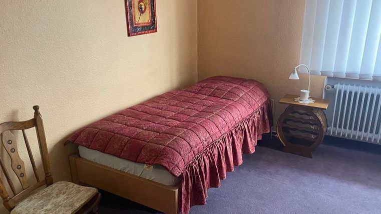 Ein einfaches Zimmer mit einem Einzelbett und einer roten Bettdecke. Ein Stuhl und ein Nachttisch mit einer Lampe und einem Glas befinden sich ebenfalls im Raum.