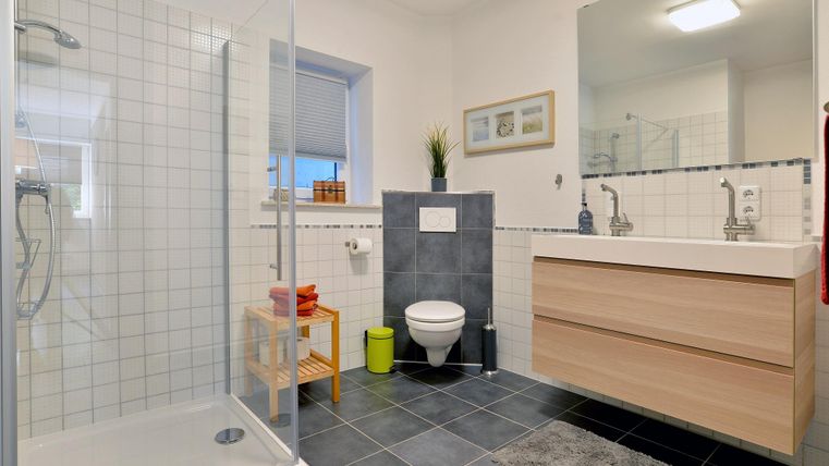 Ein modernes Badezimmer mit einer Duschkabine und einer großen Spiegelfläche. Die Möbel sind aus hellem Holz und der Boden ist mit dunklen Fliesen ausgelegt.