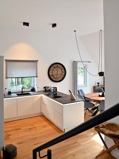 Eine moderne Küche mit hellen Möbeln und großen Fenstern. Am Tisch stehen Stühle und eine große Wanduhr ist sichtbar.