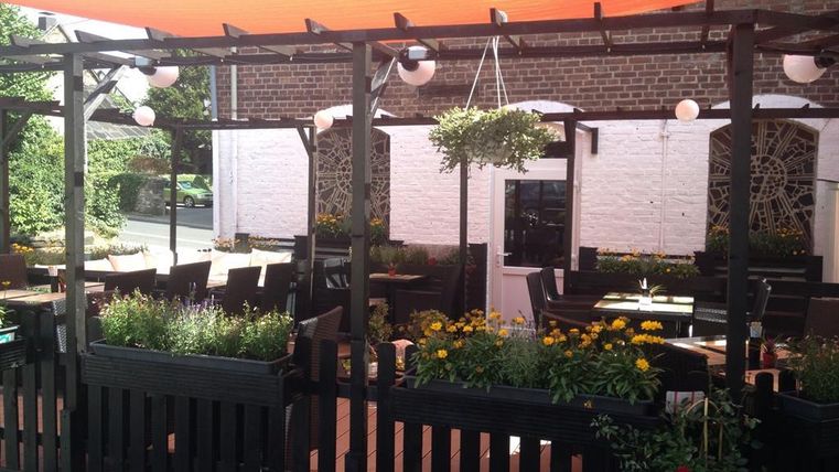 Ein gemütlicher Außenbereich eines Restaurants mit blühenden Pflanzen und Holzmöbeln. Ein rotes Sonnensegel bietet Schatten für die Gäste.