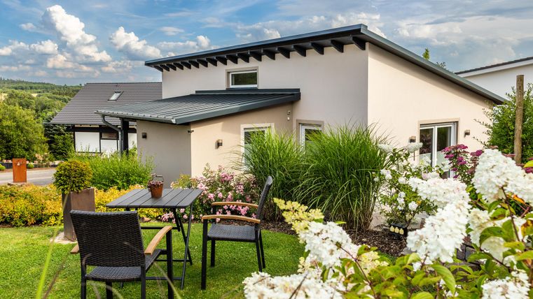 Ein modernes Haus mit einem hellen Garten. Es gibt einen Tisch mit Stühlen und Blumen umgeben von üppigem Grün.