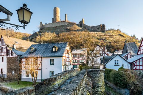 Eine malerische Landschaft mit charmanten Häusern im Vordergrund und einer alten Burg auf einem Hügel im Hintergrund. Die Szene ist von warmem Licht eines klaren Himmels beleuchtet.