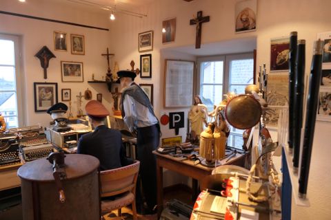 Ein nostalgisches Zimmer voller alter Erinnerungsstücke und Kunstwerke. Zwei Personen sind mit verschiedenen Gegenständen beschäftigt, umgeben von historischen Fotos und Möbeln.