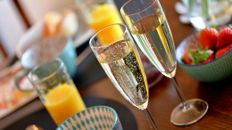 Twee glazen champagne staan op een tafel, omringd door verschillende dranken en schalen met vers fruit. Het lijkt op een feestelijk ontbijt of brunch.