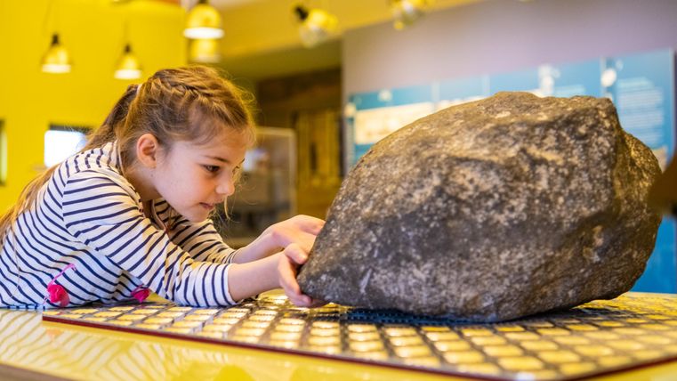 Ein kleines Mädchen versucht, einen großen Stein zu heben. Der Hintergrund zeigt eine kreative Umgebung mit bunten Wänden und interessanten Ausstellungsstücken.