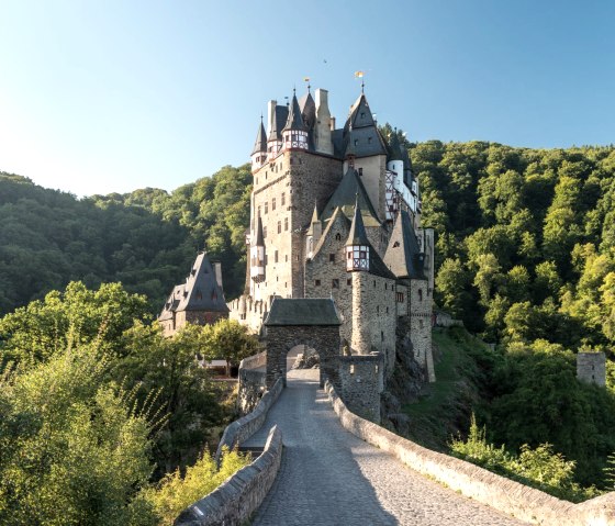 Burg Eltz thront majestätisch über einer Steinbrücke, umgeben von dichtem, grünem Wald unter klarem, blauem Himmel., © Rheinland-Pfalz Tourismus GmbH, D. Ketz
