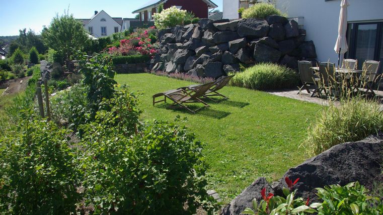 Ein einladender Garten mit einer Liege und üppigem Grün. Im Hintergrund sind Blumen und eine stilvolle Gebäudefassade zu sehen.