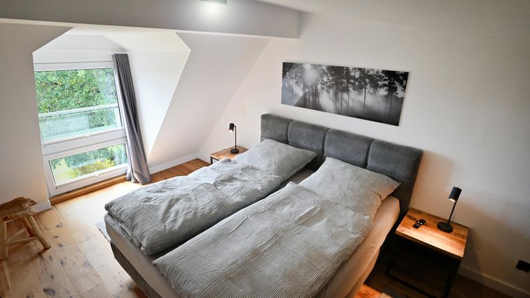Ein modernes Schlafzimmer mit einem Doppelbett und einer eleganten Bettdecke. Große Fenster sorgen für viel Tageslicht und einen Blick ins Grüne.