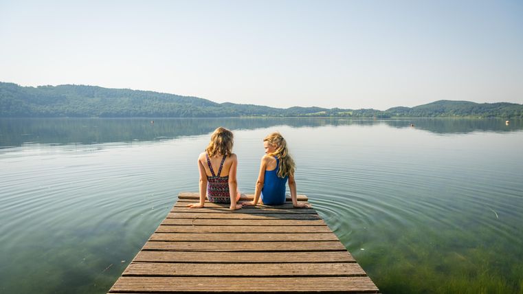 Zwei Mädchen sitzen auf einem Holzsteg am See. Im Hintergrund sind sanfte Hügel und eine ruhige Wasseroberfläche zu sehen.