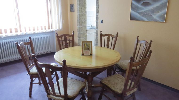 Ein gemütlicher Essbereich mit einem runden Holztisch und sechs Stühlen. An der Wand hängt ein Bild, und es gibt viel Tageslicht durch die Fenster.