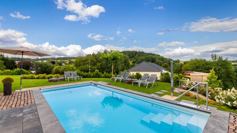 Ein schöner Pool umgeben von grünen Wiesen und Bäumen. Im Hintergrund sind sanfte Hügel und ein blauer Himmel mit weißen Wolken zu sehen.
