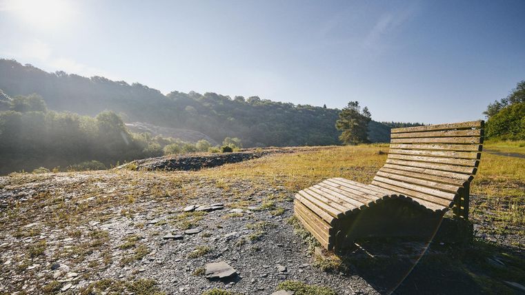 Eine Holzbank steht in einer ruhigen Landschaft. Im Hintergrund sind sanfte Hügel und ein klarer Himmel zu sehen.