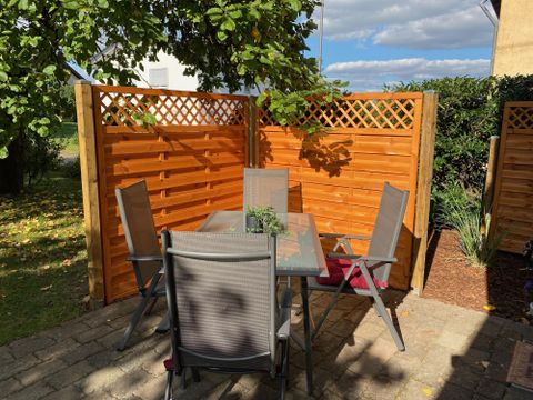 Ein gemütlicher Gartenbereich mit einem Tisch und Stühlen aus Metall. Die Holzwände sorgen für Privatsphäre und der Himmel ist teils bewölkt.