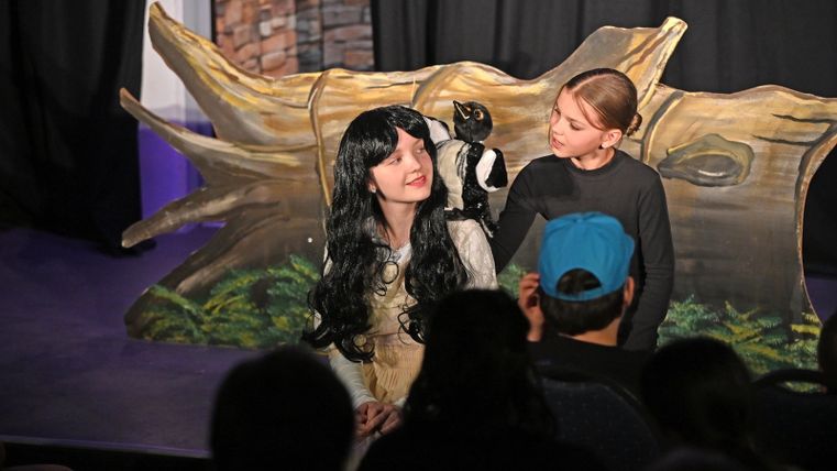 Kinder des Theaterverein Kaisersesch spielen auf einer kleinen Bühne eine Szene aus „Schneewittchen und die 7 Zwerge“: Ein Mädchen mit schwarzer Perücke sitzt vor einem Baumstamm-Bühnenbild, während ein anderes Kind mit einer Handpuppe spricht; im Vordergrund schaut das Publikum zu.