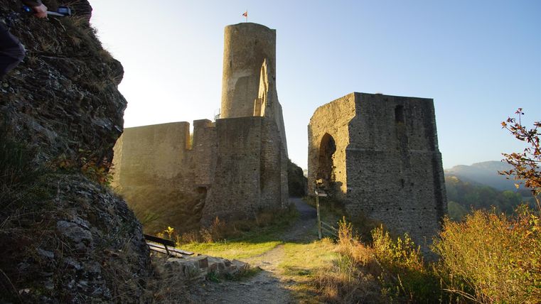Eine alte Burruine mit einem Turm und Mauern, umgeben von grünen Wiesen. Die Sonne scheint und schafft eine malerische Atmosphäre.