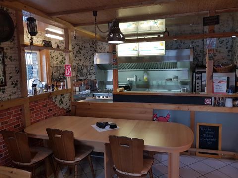 Eine gemütliche Holzstube mit einem großen Tisch und Stühlen. Im Hintergrund befindet sich eine offene Küche mit Blick auf den Grillbereich.