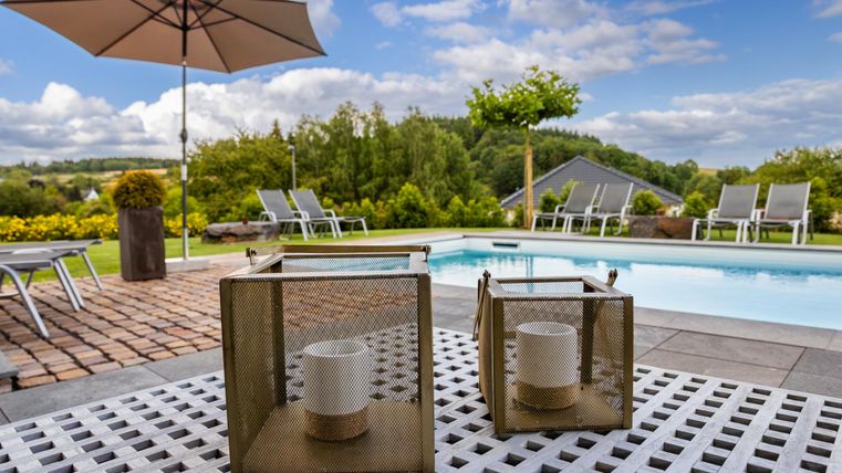 Ein schöner Poolbereich mit Liegen und einem Sonnenschirm. Auf einem Sichtschutz stehen zwei laternenähnliche Objekte.