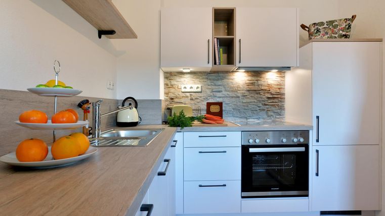 Een moderne keuken met witte meubels en een houten bord. Op een tafel staan verse vruchten, en de keuken is goed verlicht.