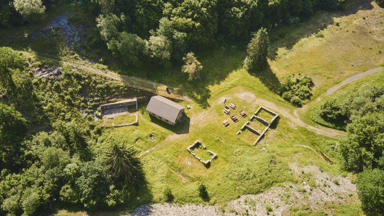 Eine Luftaufnahme eines grünen Gebiets mit einem kleinen Haus und Überresten von Gebäuden. Umgeben von Bäumen und Wiese ist die Landschaft sehr idyllisch.